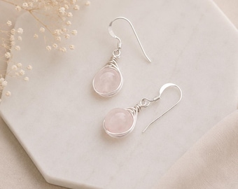 Puede incluir: Un par de pendientes colgantes con piedras preciosas redondas de cuarzo rosa pálido. Cada piedra está envuelta en un fino alambre de color plateado y unida a un sencillo gancho de color plateado para un aspecto limpio y equilibrado.