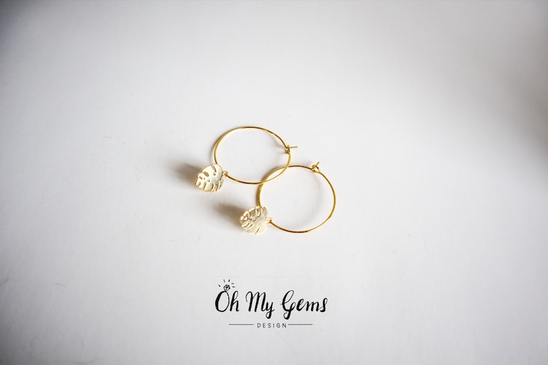 May include: A pair of gold hoop earrings featuring a small, textured monstera leaf charm attached to each hoop. The text "Oh My Gems DESIGN" is printed in black below the earrings, with a small diamond icon above the word Oh.