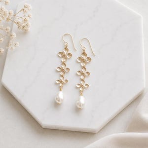 May include: A pair of gold-toned drop earrings featuring a vertical arrangement of three small, textured floral-shaped metal components, finished with a single white teardrop-shaped pearl dangling from the base of each earring on a hook wire.