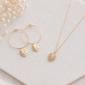 May include: A gold jewelry set consisting of two hoop earrings and a necklace. Each earring features a small, dangling monstera leaf charm. The necklace includes a delicate gold chain with a single monstera leaf pendant encrusted with small clear stones.