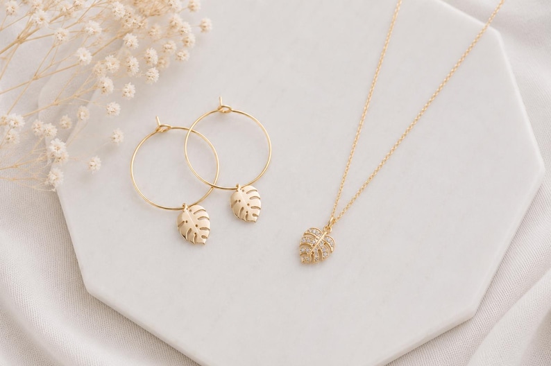 May include: A gold jewelry set consisting of two hoop earrings and a necklace. Each earring features a small, dangling monstera leaf charm. The necklace includes a delicate gold chain with a single monstera leaf pendant encrusted with small clear stones.