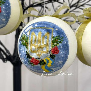 May include: Embroidered Christmas ornament with a blue and white design featuring the Ukrainian coat of arms, a wreath, and red berries. The ornament has a gold ribbon for hanging. The text "Liena_ukraina" is visible.
