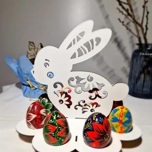 May include: White wooden bunny-shaped Easter egg holder with intricate floral designs. The holder has four slots for displaying painted Easter eggs. The eggs are decorated with colorful floral patterns.