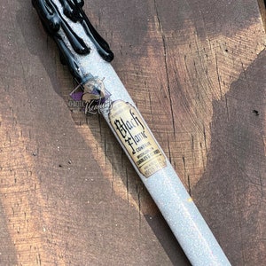 May include: A black and white pen with a black wax candle design. The pen has a silver clip and a white glitter body. The pen has a label that says "Black Flame Company Specializing in Candles & Curses"