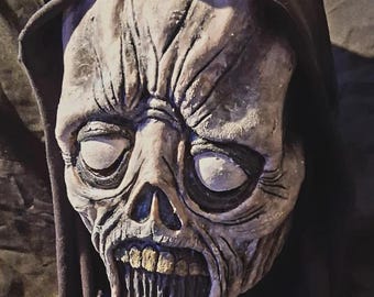 May include: A detailed, realistic, white and grey zombie mask with a black hooded robe. The mask has a wide open mouth with visible teeth and large, white eyes.