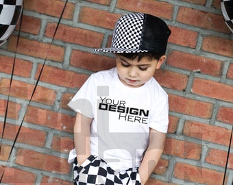 Puede incluir: Un niño con una camiseta blanca con el texto "YOUR DESIGN HERE", una gorra de béisbol a cuadros blancos y negros y pantalones cortos a cuadros. El fondo es una pared de ladrillo rojo.