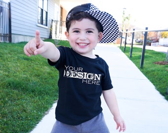 Puede incluir: Un niño sonriente lleva una camiseta negra con el texto "YOUR DESIGN HERE" en una fuente blanca desgastada. El niño también lleva una gorra de béisbol a cuadros en blanco y negro. El niño está señalando con su dedo índice derecho.