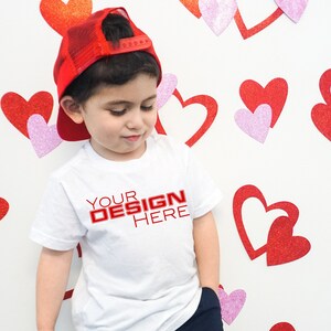 Puede incluir: Un joven lleva una camiseta blanca con el texto "YOUR DESIGN HERE" en rojo. Tambi&eacute;n lleva una gorra de camionero roja. El fondo presenta decoraciones de corazones rojos y rosas.