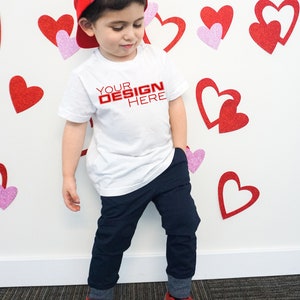 Puede incluir: Un ni&ntilde;o con una camiseta blanca con el texto "YOUR DESIGN HERE" en rojo, pantalones azul marino, una gorra de b&eacute;isbol a cuadros rojos y zapatillas rojas. El fondo presenta decoraciones de corazones rojos y rosas.