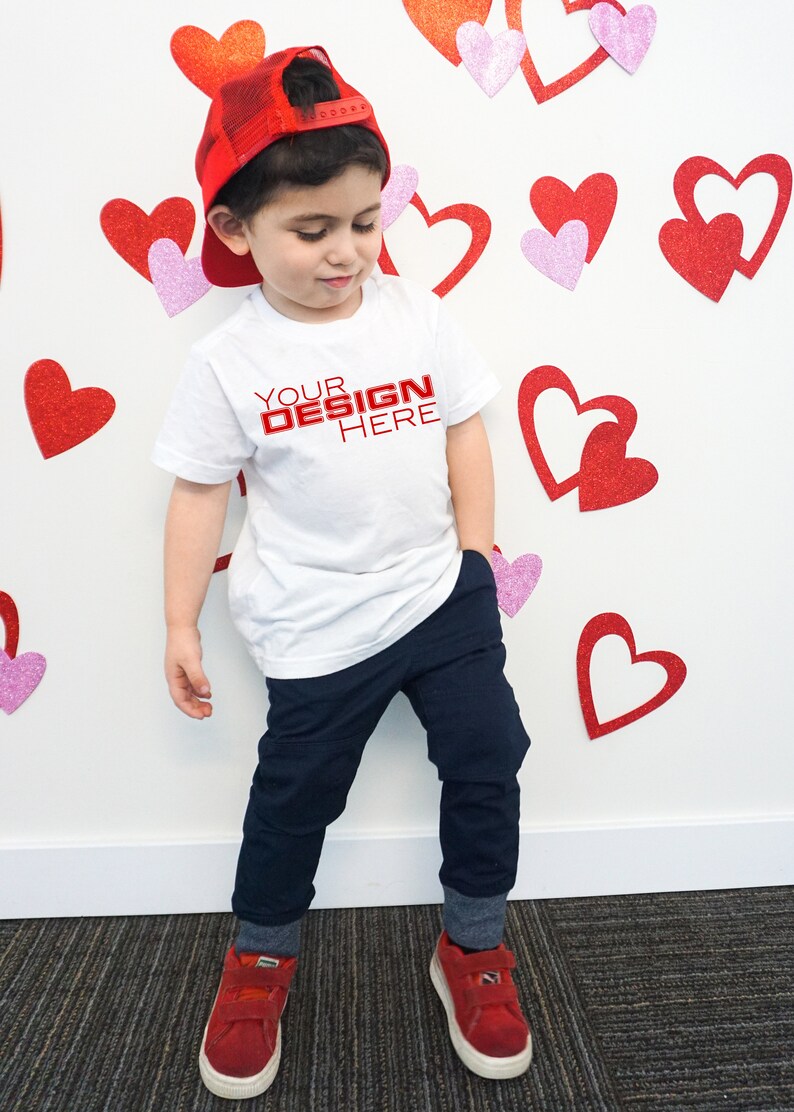 Puede incluir: Un ni&ntilde;o con una camiseta blanca con el texto "YOUR DESIGN HERE" en rojo, pantalones azul marino, una gorra de b&eacute;isbol a cuadros rojos y zapatillas rojas. El fondo presenta decoraciones de corazones rojos y rosas.