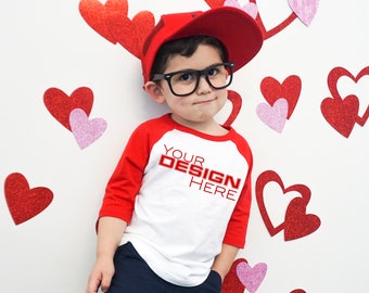 Puede incluir: Un niño con una gorra de béisbol roja, gafas negras y una camiseta raglán blanca y roja con el texto "YOUR DESIGN HERE". El fondo presenta decoraciones de corazones rojos y rosas.