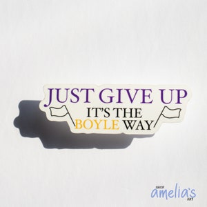 May include: A white sticker with the text "JUST GIVE UP IT'S THE BOYLE WAY" in purple and yellow. The sticker has two small flag illustrations and the shop name "amelia's art" in the bottom right corner.