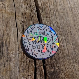 May include: A circular holographic sticker featuring a black line drawing of a bug with the text "queer bug" centred above and below the insect. The design is surrounded by small stars, hearts, and dots, with a reflective rainbow finish.