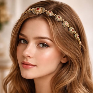 Może przedstawiać: Złota opaska do włosów z czerwonymi i przezroczystymi kamieniami szlachetnymi. Ozdobna ozdoba do włosów jest noszona na falowanych, jasnobrązowych włosach. Obraz to zbliżenie osoby o jasnej karnacji i miękkim, naturalnym makijażu.