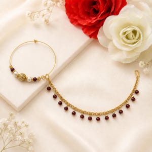 May include: A gold nose ring with a chain and red beads. The nose ring has a circular design with pearls and a decorative gold bead. The chain is decorated with small, round, red beads, and is attached to the nose ring.