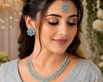 May include: A jewelry set consisting of a silver-toned necklace, matching stud earrings, and a maang tikka headpiece. Each piece features a floral-shaped design with bright turquoise stones and clear crystals set in a metallic silver-toned frame.