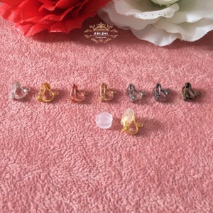 May include: A collection of clip-on earrings in various metallic finishes, including silver, gold, and bronze, arranged on a pink textured surface. The earrings have different shapes and designs, with a clear, round earring back and a gold-toned clip-on earring.