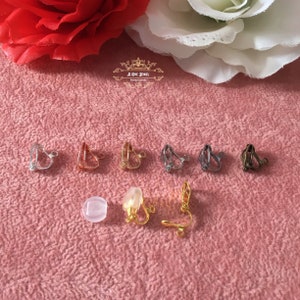May include: A collection of clip-on earring findings in various metallic finishes, including silver, gold, and bronze. The image also shows a clear, round earring pad and a gold-colored earring clip. The background is a textured pink surface with floral accents.