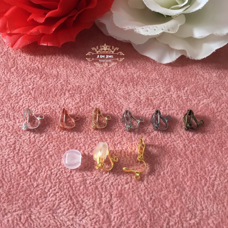 May include: A collection of clip-on earring findings in various metallic finishes, including silver, gold, and bronze. The image also shows a clear, round earring pad and a gold-colored earring clip. The background is a textured pink surface with floral accents.
