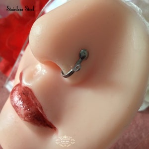 May include: A stainless steel nose ring is shown on a mannequin. The nose ring is silver and has a small, circular design. The text "Stainless Steel" is visible in the upper left corner.