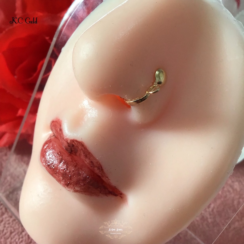 May include: A gold-colored nose ring is displayed on a mannequin head. The nose ring is a small, delicate piece of jewelry. The mannequin has red lipstick and the text "KC Gold" is visible in the upper left corner.