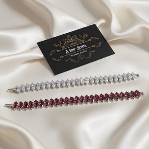 Könnte beinhalten: Zwei elegante Armbänder mit Marquise-Schliff-Edelsteinen. Eines hat klare Steine, das andere tiefrote Steine. Die Armbänder werden auf einer Satin-Oberfläche präsentiert, mit einer Visitenkarte "A One Jewel Premium Jewelry."