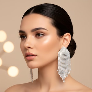 May include: A woman wearing large, dangling, silver rhinestone earrings. The earrings are rectangular at the top and cascade into multiple strands of rhinestones, creating a shimmering, elegant effect. The background is a soft, neutral color.