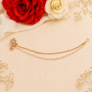 May include: A gold-coloured nose chain with a floral design and dangling pearls. The chain features two strands of pearls and a delicate hook. Red and white roses and small white flowers are in the background.