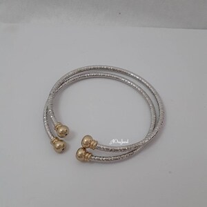 May include: Two silver bangle bracelets with gold accents. The bracelets are made of a textured metal and have a rounded shape.