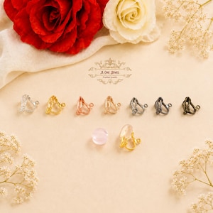 May include: A collection of clip-on earrings in various metallic finishes, including silver, gold, and bronze, arranged on a cream-colored surface. A clear and a pink gemstone are also present. The image is set against a backdrop of red and white roses.