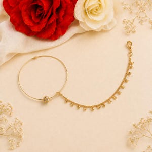 May include: A gold nose ring featuring a large hoop and a decorative ball. A delicate chain with small gold beads is attached to the ball. The jewellery is set against a cream background with red and white roses.