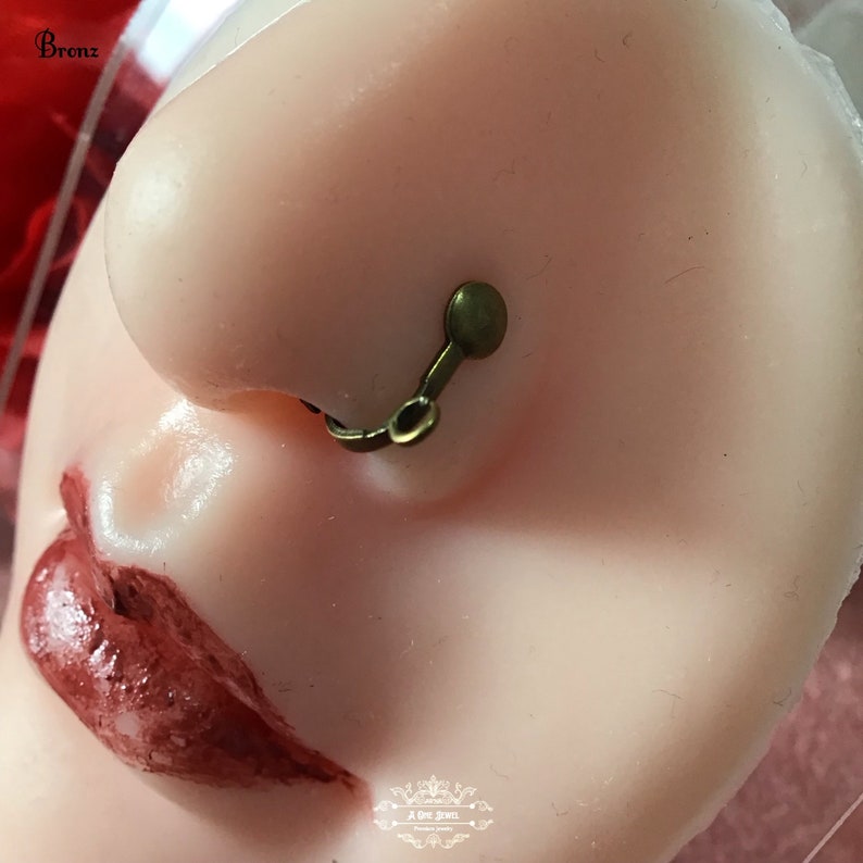 May include: A close-up of a nose with a bronze-colored, non-piercing nose ring. The nose ring has a small disc shape on the top and a curved design that connects to the nostril. The skin tone is light.
