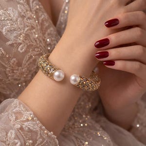 May include: A gold-coloured cuff bracelet featuring a pearl at each end. The bracelet has an intricate, textured pattern. The person's nails are painted a deep red. The background is a soft, neutral colour, suggesting a formal event.