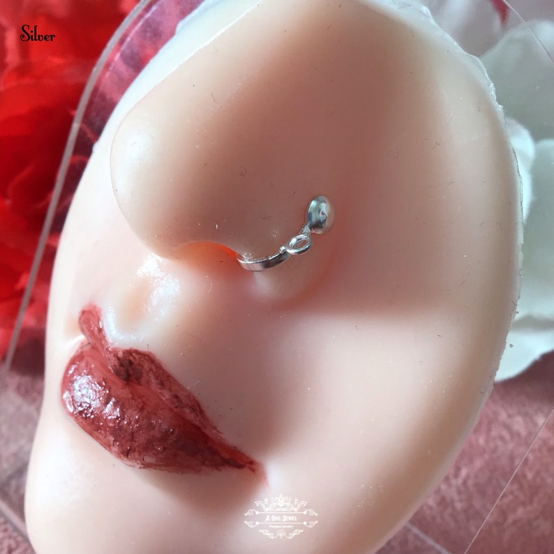 May include: A silver nose ring with a small dangling charm. The nose ring is attached to the nostril with a small hoop and the charm hangs below. The word "Silver" is in the top left corner.