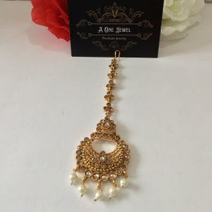 May include: Gold-toned headpiece with a teardrop-shaped pendant and a crescent-shaped design with white pearls hanging from the bottom. The headpiece is adorned with clear rhinestones.