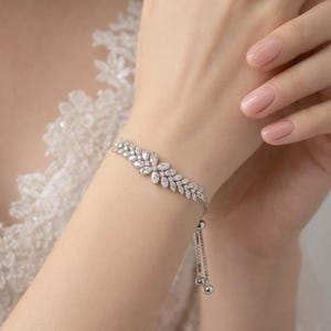 Op de afbeelding: Een zilveren armband met een bloemmotief. De armband heeft een reeks heldere, sprankelende stenen gerangschikt in een bladvormig patroon. De armband is verstelbaar met een delicate ketting en twee kleine zilveren ballen.