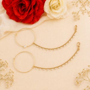 Op de afbeelding: Twee goudkleurige neusringen met een parelaccent en een delicate ketting versierd met kleine parels. Op een crèmekleurige achtergrond, met rode en witte rozen. Een prachtig sieraad.