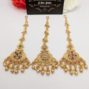 May include: Three gold-colored headpieces with intricate designs. Each piece features a central floral motif with red, green, and clear gemstones, and dangling gold beads. The headpieces are displayed on a white surface with a business card in the background.