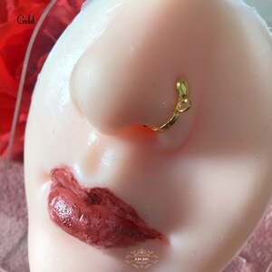 May include: A close-up of a gold-colored nose ring. The nose ring is a simple, curved design with a small loop at the end. The word "Gold" is in the upper left corner. The background is a light skin tone.