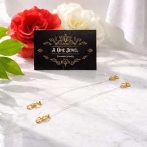May include: Two clear, thin jewelry straps with gold-colored clasps are displayed. A black card with gold text that reads "A One Jewel Premium Jewelry" is behind the straps. Red and white roses are in the background.