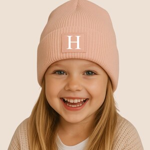Könnte beinhalten: Hellrosa Beanie mit weißem Buchstaben "H" auf einem rechteckigen Aufnäher. Die gerippte Strickmütze wird von einer jungen Person mit blonden Haaren und einem lächelnden Gesicht getragen.