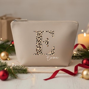Könnte beinhalten: Beige Kosmetiktasche mit Reißverschluss, mit einem großen Leopardenmuster-Buchstaben "E" und dem Namen "Emma". Weihnachtsdekorationen, darunter Kugeln und ein rotes Band, umgeben die Tasche.