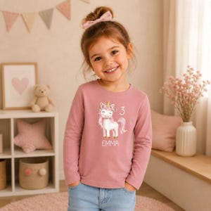 Könnte beinhalten: Ein langärmeliges, staubrosafarbenes Shirt mit einem weißen Einhorn-Motiv mit Blumenkrone, der Zahl 3 und dem Namen "EMMA". Das Shirt wird mit einer blauen Jeans kombiniert.