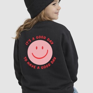 Könnte beinhalten: Schwarzes Sweatshirt mit einem rosa Smiley-Gesicht und dem Text "It's a good day to have a good day" in Rot. Die Person auf dem Bild trägt eine schwarze Mütze.