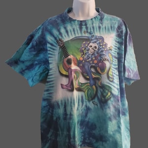 May include: A tie-dye t-shirt with a colorful graphic of a skull wearing a jester's hat and holding a guitar. The graphic is surrounded by a swirl of colors.