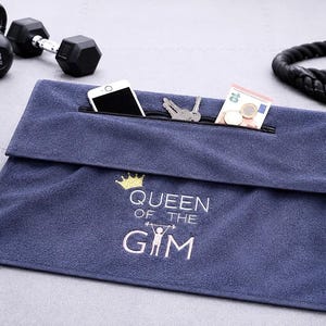 Könnte beinhalten: Ein rechteckiges, marineblaues Sporthandtuch mit einer Reißverschlusstasche an der Oberkante. Das Handtuch ist mit einer goldenen Krone über dem weißen Schriftzug "QUEEN OF THE GYM" bestickt, wobei das "I" durch eine Hanteln hebende Figur ersetzt ist.