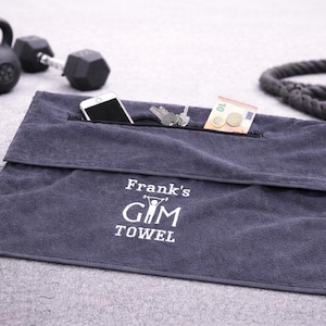 Op de afbeelding: Een donkergrijze rechthoekige sporthanddoek met een opbergvak met ritssluiting langs de bovenrand. De handdoek is geborduurd met de witte tekst "Frank's GYM TOWEL" en een kleine afbeelding van een persoon die een halter optilt.