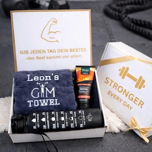 Könnte beinhalten: Eine weiße Geschenkbox mit einem dunkelgrauen Handtuch mit der Stickerei Leon's Gym Towel, einer Tube Kneipp Men Energie Duschgel und einer schwarzen Trinkflasche mit stündlichen Markierungen. Der Deckel zeigt einen goldenen Bizeps und deutschen Text.