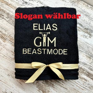 Op de afbeelding: Zwarte gymhanddoek met goudkleurige geborduurde tekst: "ELIAS", "GIM" en "BEASTMODE". Een gouden lint is om de handdoek gebonden. De tekst "Slogan wählbar" staat in het rood bovenaan.