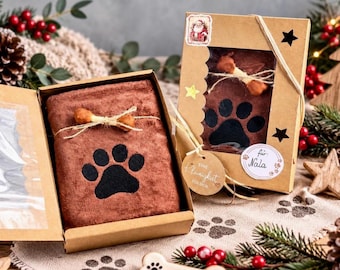 Könnte beinhalten: Ein braunes Handtuch mit schwarzem Pfotenabdruck, verpackt in einer braunen Kartonschachtel. Die Box hat ein Sichtfenster, einen Weihnachtsmann-Aufkleber, Stern-Ausstanzungen und ein rundes Etikett mit der Aufschrift "für: Nala" und "Eine Kleinigkeit für dich."
